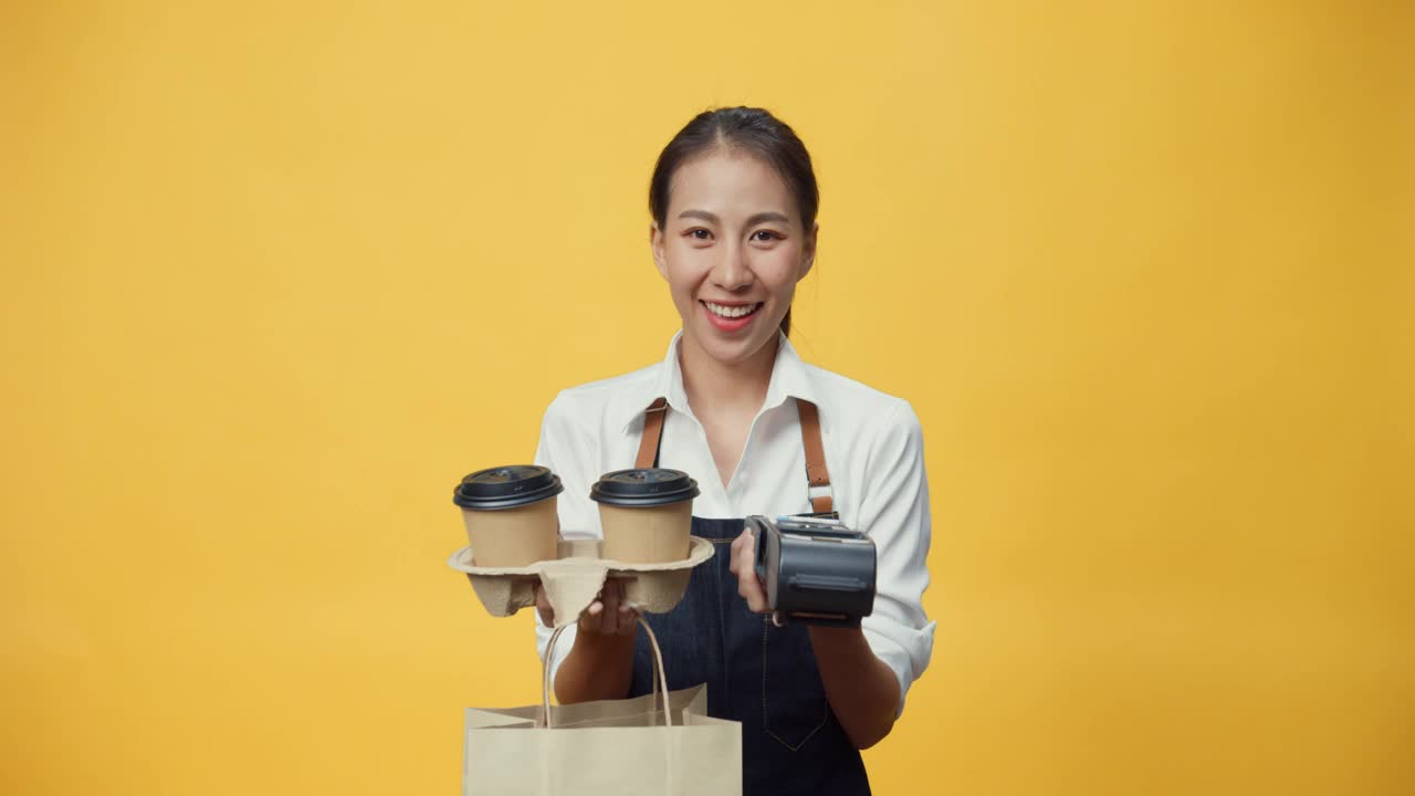 快乐微笑的亚洲女服务员穿着围裙友好地看着相机给顾客咖啡或茶和信用卡读卡机孤立在黄色背景。