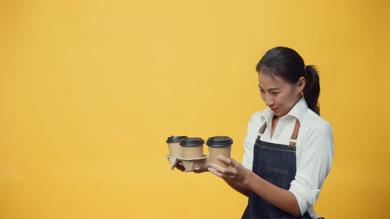 快乐微笑的亚洲女士企业老板穿着围裙友好地看广告文案空间持有咖啡或茶孤立在黄色的背景。