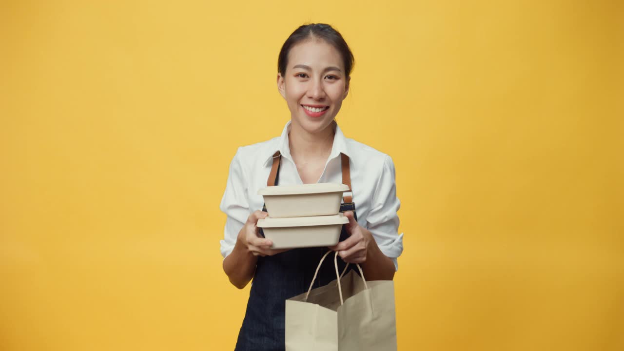 快乐微笑的亚洲女士商人友好地看着相机美味的食品和饮料放在纸盒里的工艺纸袋孤立在黄色的背景。