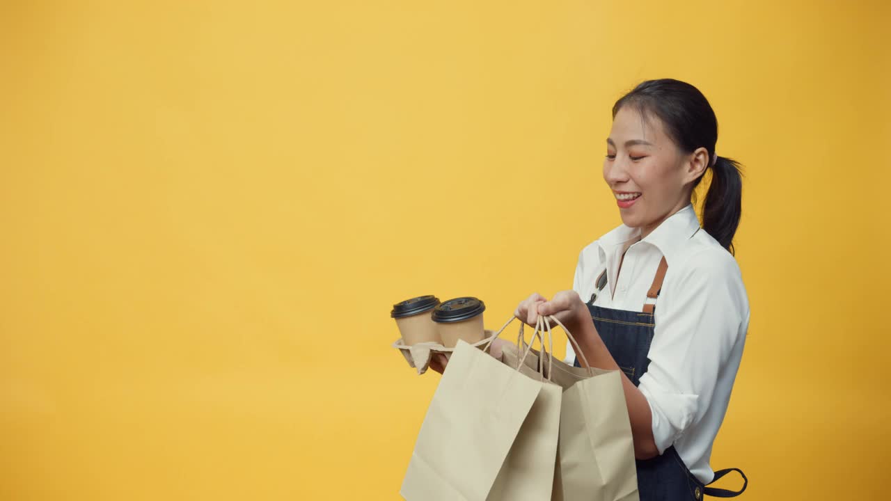 快乐微笑的亚洲女服务员，穿着围裙，友好地看着文案空间的广告，装着咖啡或茶和美味的食物在手工纸袋隔离在黄色的背景上。