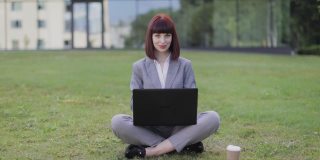 穿着正装的年轻女子，盘腿坐在绿草地上，在办公室外使用笔记本电脑