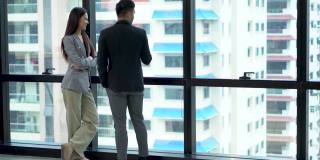 快乐的亚洲商人和女商人讨论商业战略在一个高楼大厦的现代办公室城市景观的背景。两个工人在城市摩天大楼的窗前交谈