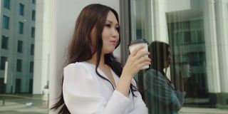 年轻英俊的亚洲女商人正在喝咖啡的背景下，她的反思在商业中心。夏季白色商业区