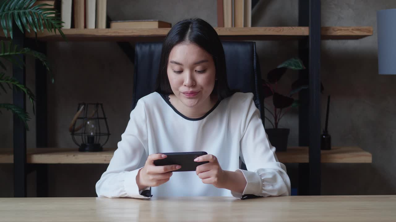 一个情绪化的年轻亚洲女人正坐在公寓的桌子上玩她的智能手机游戏