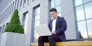 男性亚洲商人工作与笔记本电脑在线户外坐在长椅在市中心的城市