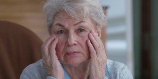 资深女性抚摸着布满皱纹的脸，表情严肃地看着镜头。焦虑悲伤的白人女性退休在家的头像。老化的概念