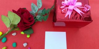 一个礼品盒系蝴蝶结丝带和红玫瑰和一张空白贺卡在红色背景的桌子上。平的。直接上图。设计情人节，周年纪念或节日庆祝装饰