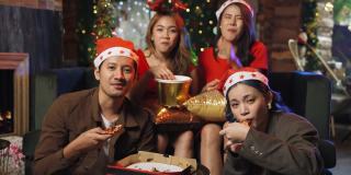 一群亚洲年轻人戴着圣诞帽，一边看电视，一边吃着爆米花和披萨