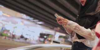 两名穆斯林妇女戴着头巾站在路边，用手机呼叫出租车取车服务，城市夜间公共交通移动应用方便便捷的城市城市智能生活