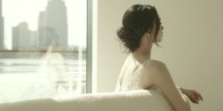 身穿白色婚纱的美丽亚洲新娘坐在落地窗前的沙发上慢镜头拍摄