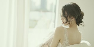 身穿白色婚纱的美丽亚洲新娘坐在落地窗前的沙发上慢镜头拍摄