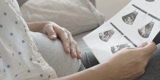 亚洲孕妇看超声波图像和触摸她的腹部坐在沙发上。