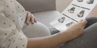 亚洲孕妇看超声波图像和触摸她的腹部坐在沙发上。
