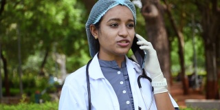 穿着白大褂的印度治疗师，脖子上用听诊器取下面罩，用手机与患者交谈。使用现代技术远程提供帮助