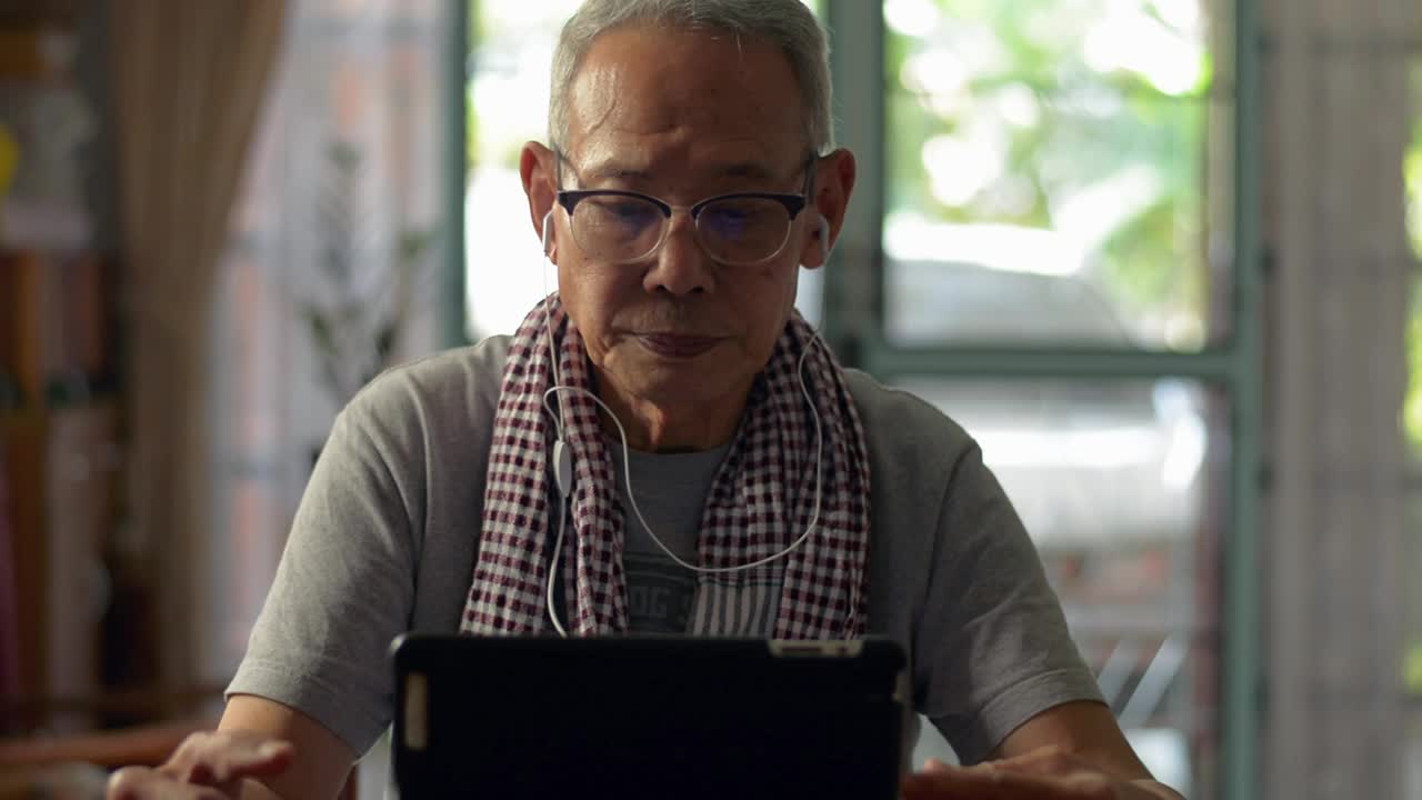一位戴着眼镜、戴着耳机的亚洲老人在家里的桌子上用数码平板电脑。穿着休闲装的退休老人在数码设备上观看在线社交视频。回家隔离。老年人的生活方式。