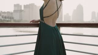 一位身穿绿色晚礼服的年轻亚洲新娘，站在湖滨码头，以城市建筑为背景，微笑着看着镜头，用慢镜头拍摄视频素材模板下载