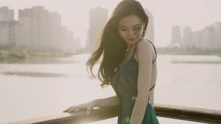 一位身穿绿色晚礼服的年轻亚洲新娘，站在湖滨码头，以城市建筑为背景，微笑着看着镜头，用慢镜头拍摄视频素材模板下载