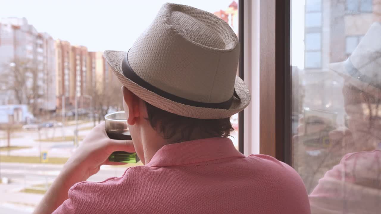 一个戴着帽子的人站在窗口，看着街道，喝着玻璃杯里的饮料