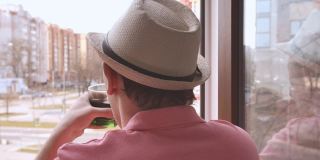 一个戴着帽子的人站在窗口，看着街道，喝着玻璃杯里的饮料