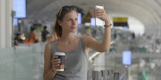 年轻的白人女性旅客在机场自拍。拿着手机和咖啡纸杯