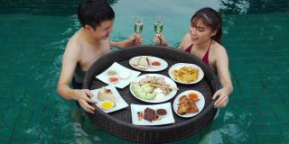 幸福的年轻夫妇享受着漂浮的食物，在游泳池里喝着香槟杯