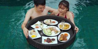 快乐的年轻夫妇享受和吃漂浮在游泳池的食物