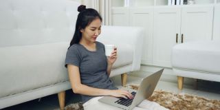 一位亚洲妇女坐在客厅里，手里拿着一个咖啡杯，膝上放着一台笔记本电脑。
