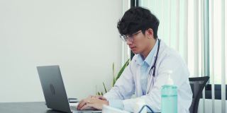 亚洲医生戴着眼镜，穿着白色制服，戴着听诊器，在新冠肺炎疫情期间向患者提供在线咨询，并保持社交距离。