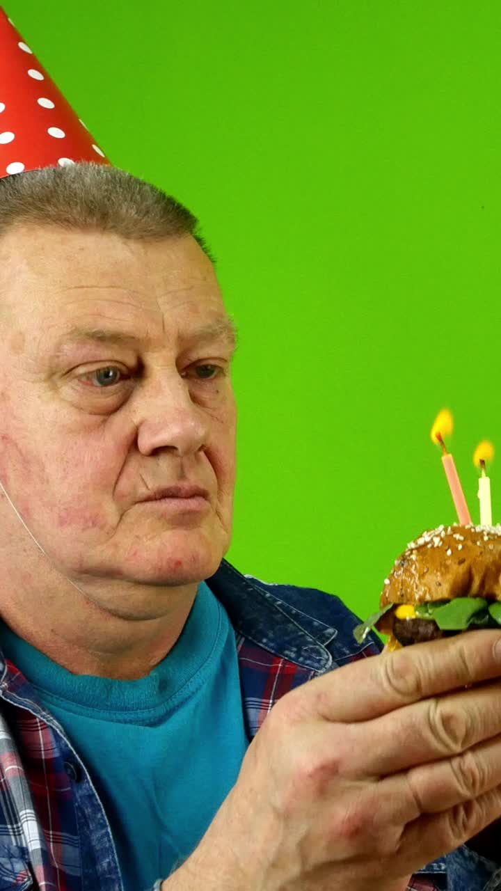 戴着生日帽的白种男性老人吹灭了生日蛋糕上的蜡烛，兴高采烈地吃着汉堡。
