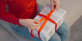 闭上女人的手用丝带打蝴蝶结为一个包装好的礼品盒。漂亮的女性穿着红衣服坐在客厅的沙发上，准备圣诞礼物庆祝圣诞传统派对