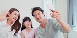 亚洲父母带着小女儿与家人在线视频通话。慈爱的爸爸、妈妈和小女孩坐在卧室的床上，周末在家里用手机和亲戚聊天。