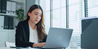 亚洲美丽忙碌的商业女性员工在办公室的桌子上工作。漂亮的职业女性穿着正式的服装坐在办公桌旁，用笔记本电脑和电话与同事交谈。