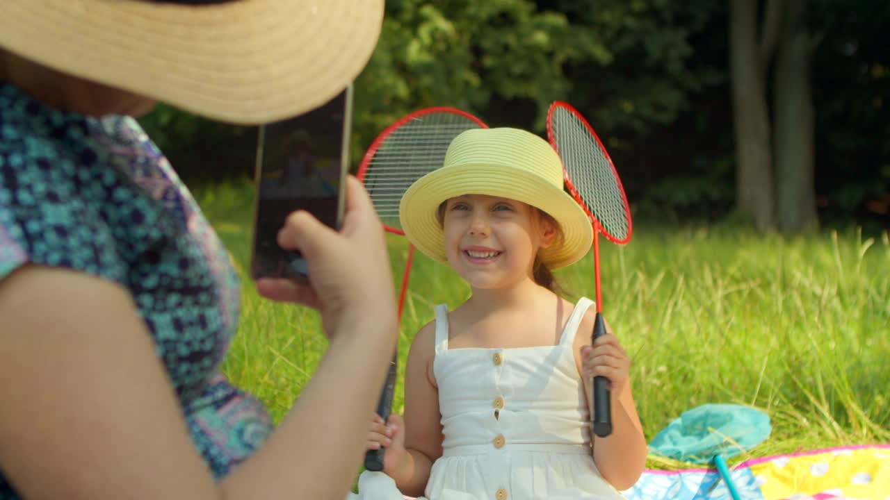 一个小女孩在她妈妈旁边用手机拍照的特写镜头。母亲用手机给坐在乡下公园里的小女儿拍了一张照片。从女人的后面看。