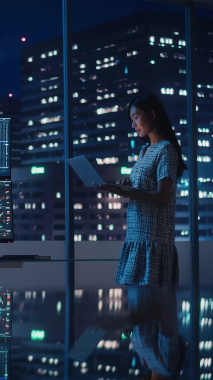 金融分析师的垂直肖像使用笔记本电脑，站在窗口与市中心街道摩天大楼霓虹灯。在投资银行城市办公室工作的女商人。