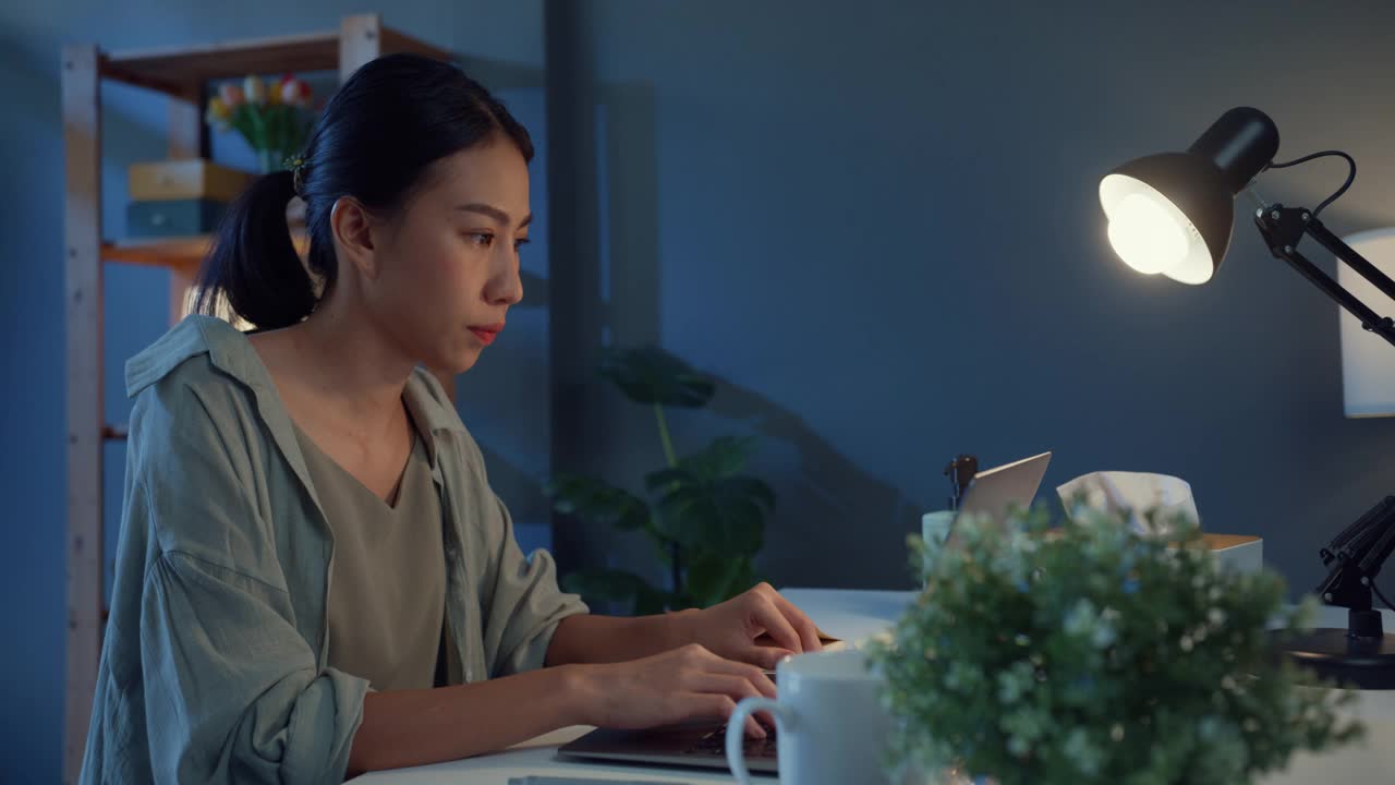 年轻的自由职业者亚洲妇女使用笔记本电脑和喝咖啡在家里的客厅晚上。