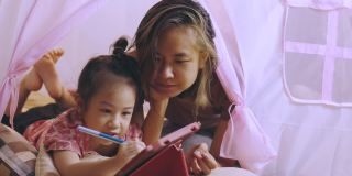 一张专注于亚洲妈妈和可爱的女儿躺在家里粉色帐篷的地板上，在使用数字技术的平板电脑上写着快乐和快乐的时刻。