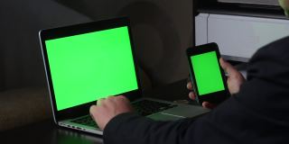 一名男子在一台绿色屏幕的笔记本电脑前工作。他手里拿着一部绿色屏幕的智能手机。附近有一枚加密货币。Chromakey。