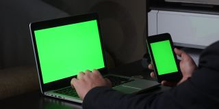 一名男子在一台绿色屏幕的笔记本电脑前工作。他手里拿着一部绿色屏幕的智能手机。附近有一枚加密货币。Chromakey