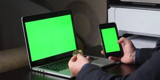 一名男子在一台绿色屏幕的笔记本电脑前工作。他手里拿着一部绿色屏幕的智能手机。考虑加密货币。Chromakey。
