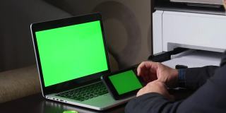 一名男子在一台绿色屏幕的笔记本电脑前工作。他手里拿着一个水平方向的绿色屏幕的智能手机。附近有一枚加密货币。Chromakey。