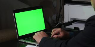 一名男子在一台绿色屏幕的笔记本电脑前工作。不时地用铅笔在纸上做笔记。Chromakey