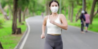 covid-19大流行期间，亚洲跑步者在公园慢跑。在冠状病毒时期锻炼。健康女性户外慢跑，健康生活理念。她脸上戴着防毒面具。