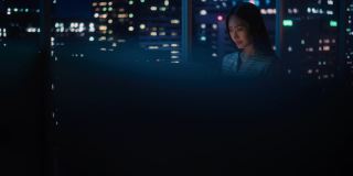 金融分析师使用笔记本电脑，站在窗户旁边与美丽的夜晚城市天际线与摩天大楼。自信的亚洲女商人在现代公司办公室工作。