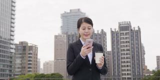 慢镜头:成功的亚洲女商人穿着西装，用着现代智能手机，在早上上班前喝着咖啡。年轻的女员工步行到办公楼。