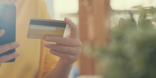 亚洲女性在家里客厅购物时，一边用手机一边用信用卡进行网上交易。