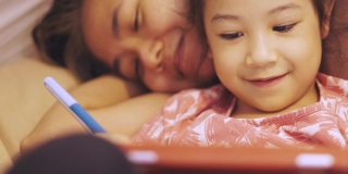 亚洲妈妈和可爱的女儿躺在家里的地板上，在周末用科技娱乐的平板电脑上书写快乐快乐的时刻。