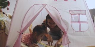 一位亚洲妈妈正在给她可爱的小女孩讲睡前故事，小女孩正躺在家里可爱的帐篷里，享受着和家人一起学习的快乐和幸福。