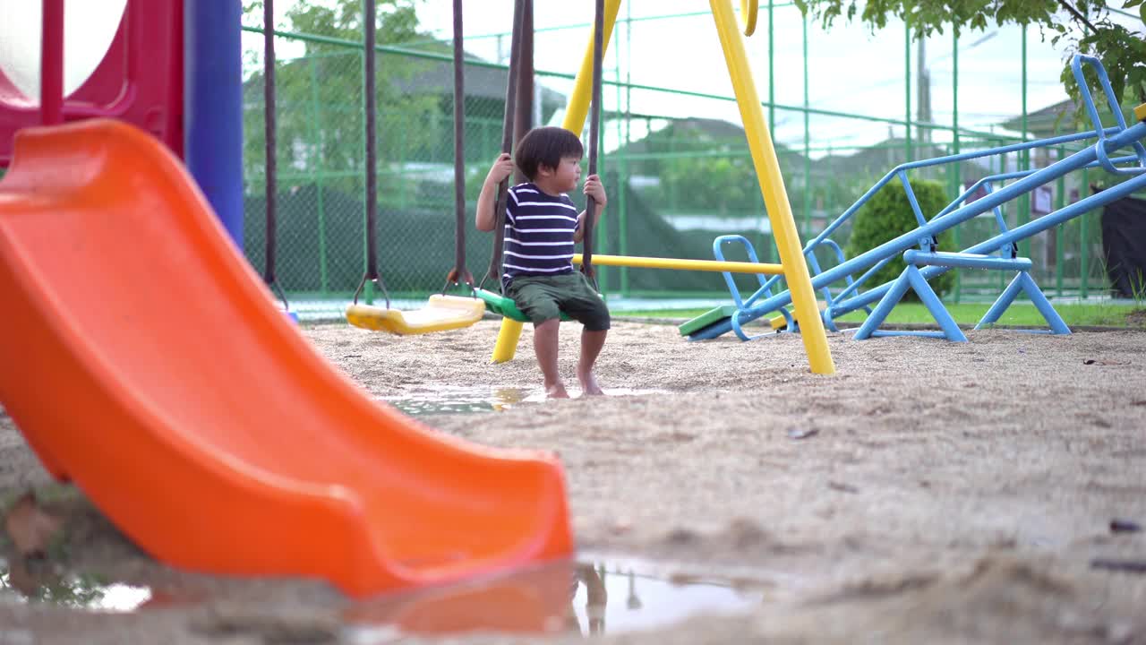 雨后的操场上，一个蹒跚学步的男孩正在玩秋千，他的脚踩在水坑上