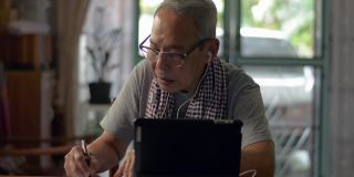 一位亚洲老爷爷穿着便装，在家用电子平板电脑上观看在线学习视频教程，并在纸上写字。老年人教育技术。