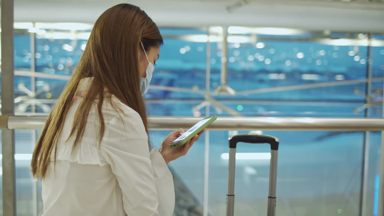 亚洲女性戴着医用口罩在机场使用智能手机，跟踪拍摄